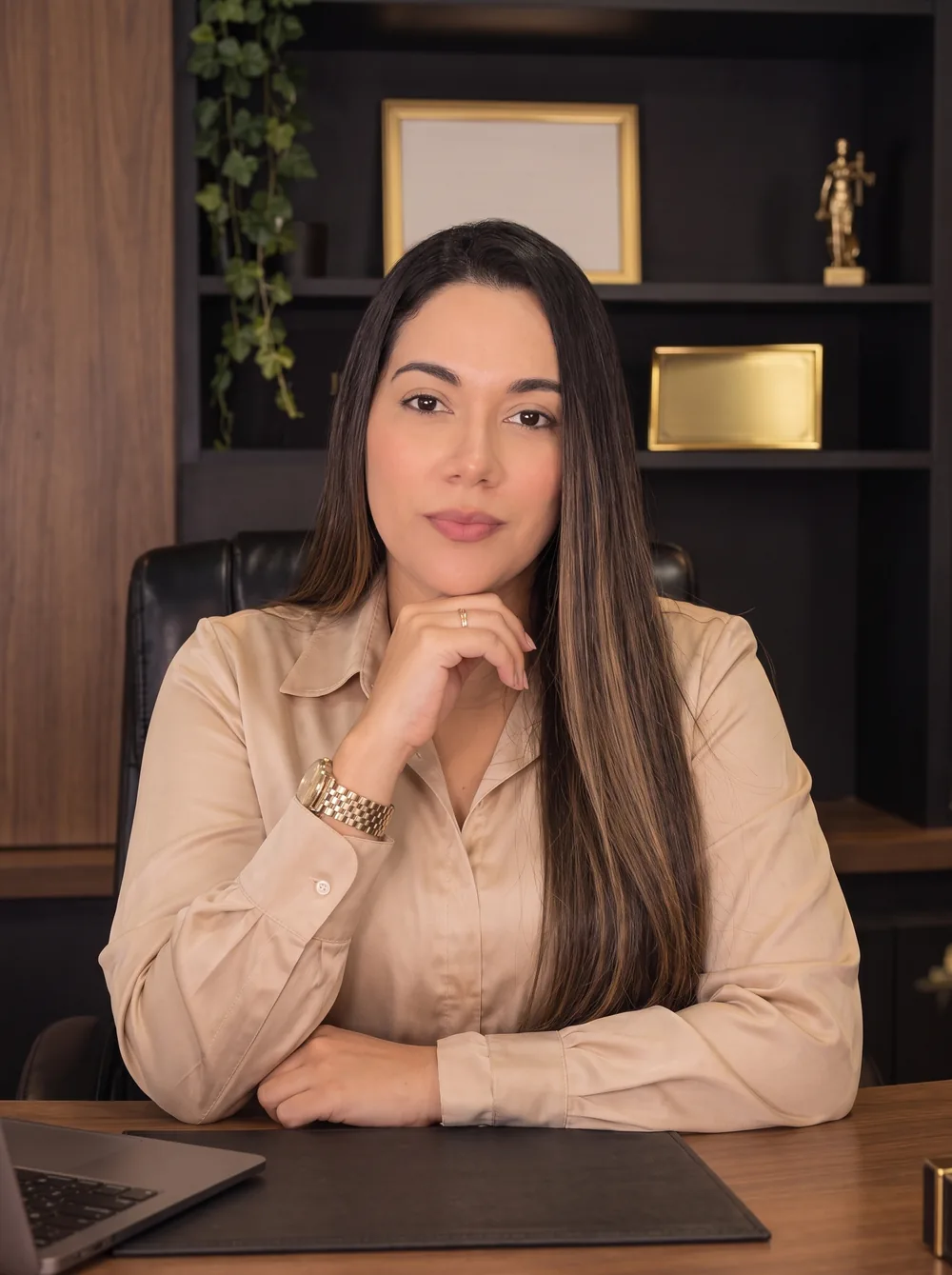 Dra. Jackeline Santos, advogada, de camisa bege e cabelo longo, com a mão apoiada no queixo, sentada à mesa do escritório.